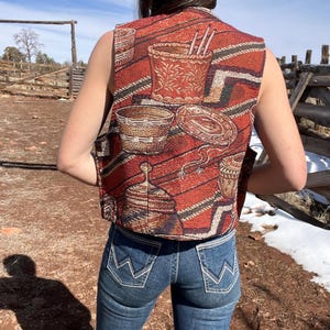 May include: A sleeveless vest with a Southwestern-inspired pattern in shades of red, brown, and black. The vest features a woven design with images of baskets and pottery. The person is wearing blue jeans.