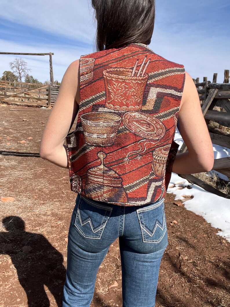 May include: A sleeveless vest with a Southwestern-inspired pattern in shades of red, brown, and black. The vest features a woven design with images of baskets and pottery. The person is wearing blue jeans.