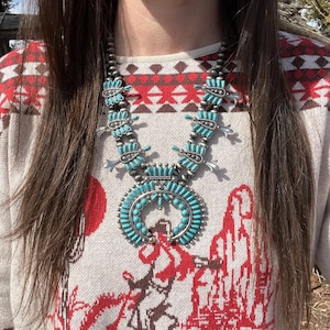 May include: A turquoise and silver Native American style necklace with a large pendant. The necklace is worn by a person wearing a white and red patterned sweater.