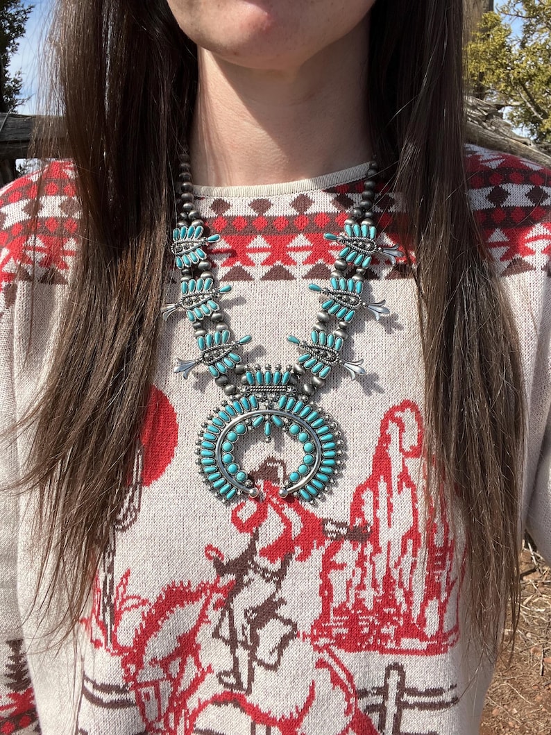 May include: A turquoise and silver Native American style necklace with a large pendant. The necklace is worn by a person wearing a white and red patterned sweater.