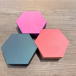 May include: Three hexagonal coasters in shades of pink, coral, and teal. The coasters are arranged on a wooden surface. The pink coaster has a textured surface, while the teal coaster has a subtle pattern.