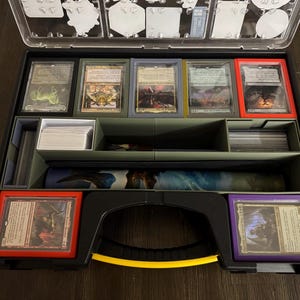 May include: A black carrying case with a yellow handle, containing a collection of trading cards. The case has multiple compartments for storing and organizing cards. The cards are visible in the top row, with a variety of colorful illustrations.