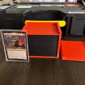 May include: A red and black card storage box with a clear plastic sleeve containing a Magic: The Gathering card featuring Purphoros, Bronze-Blooded, a Legendary Enchantment Creature - God. The card text reads "Indestructible. As long as your devotion to red is less than five, Purphoros isn't a creature.  {2}{R}: Purphoros deals 2 damage to each creature and each player.  {4}{R}{R}: You get a creature token that's a 4/4 red Elemental creature with haste.  You may cast this card only if your devotion to red is five or more. (Each {R} in the mana cost of a spell you cast represents a red mana symbol on the field.)"