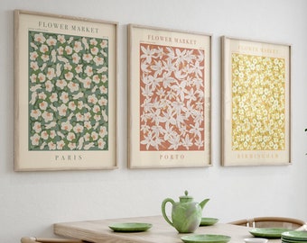May include: Three framed botanical prints with the text "Flower Market" and the city names "Paris", "Porto", and "Birmingham" hanging on a white wall above a wooden dining table with four chairs. The prints feature different floral patterns in green, orange, and yellow.