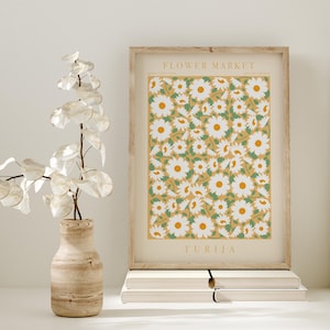 May include: A framed print with a daisy pattern on a yellow background. The print is labelled "Flower Market" and "Turija." A wooden vase with a plant and a stack of books are in the foreground.