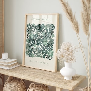 Op de afbeelding: Een ingelijste print met een botanisch bladerontwerp. Het kunstwerk toont verschillende tinten groene bladeren gerangschikt in een dicht patroon. De woorden "NATURE SPIRIT" en "LEAF COLLECTION" staan op het kunstwerk gedrukt. De lijst is van lichtgekleurd hout.