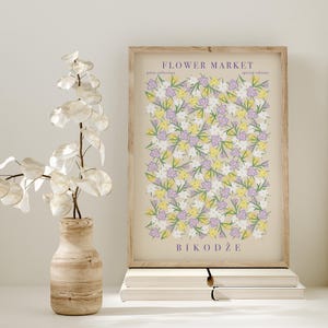 May include: A framed print with a floral design in yellow, lilac, and white, titled "Flower Market". The print is in a light wood frame, placed on a stack of white books. A decorative vase with a plant is on the left.