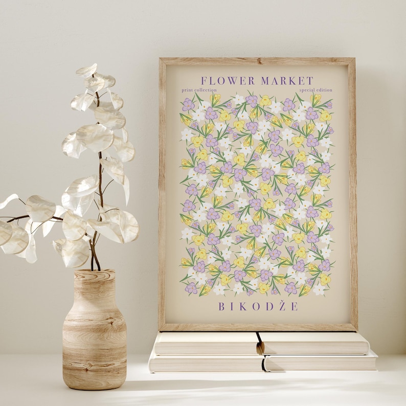 May include: A framed print with a floral design in yellow, lilac, and white, titled "Flower Market". The print is in a light wood frame, placed on a stack of white books. A decorative vase with a plant is on the left.