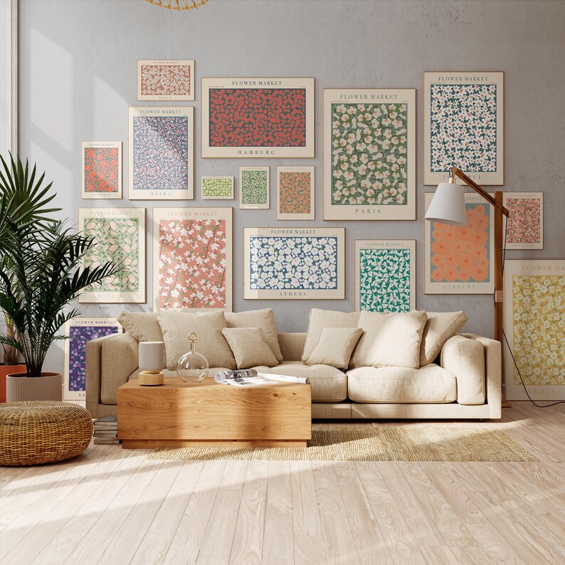 May include: A display of framed floral art prints in diverse sizes and colours, such as red, white, green, and blue, mounted on a grey wall. The prints display the text "Flower Market" and city names. A beige sofa and wooden coffee table are in the foreground.