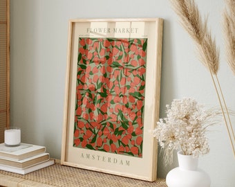 May include: A framed art print featuring a floral design with red flowers and green leaves on a light green background. The print is labeled "Flower Market Amsterdam" in a light wood frame, displayed on a woven table.