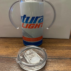 May include: A white and blue insulated tumbler with the text "Natura LIGHT" in blue and orange. The tumbler has clear handles and a clear lid. The bottom of the tumbler has a blue band with the text "America Since 1977".