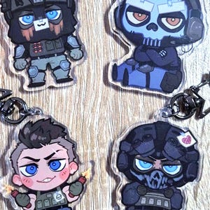Pode incluir: Quatro chaveiros de acrílico com personagens de desenhos animados em equipamento militar. Os personagens têm olhos grandes e capacetes. A paleta de cores inclui tons de azul, preto e castanho. Cada chaveiro tem um porta-chaves prateado.