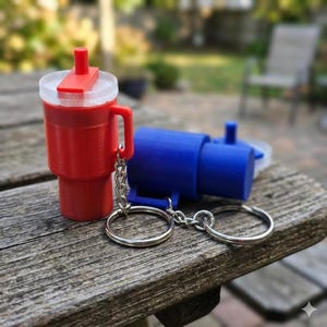 May include: A red 3D-printed tumbler with a handle and a clear lid, attached to a keychain. Beside it is a blue 3D-printed train engine, also on a keychain. Both are on a wooden surface.