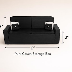 Puede incluir: Un pequeño sofá morado de dos plazas. El sofá es de color morado sólido y brillante, con un diseño minimalista. Se encuentra sobre una superficie de madera clara, posiblemente un objeto decorativo o un juguete. Sus dimensiones son de unos 15 cm de largo.