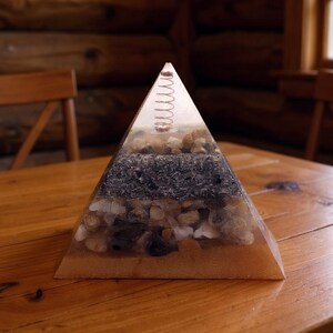 Puede incluir: Objeto decorativo en forma de pir&aacute;mide, con capas visibles de arena, piedras y un resorte en la parte superior, encapsulado en resina transparente. Colocado sobre una mesa de madera.
