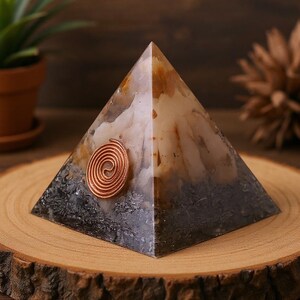 May include: A handcrafted pyramid-shaped orgone energy generator. The pyramid features a copper spiral and a mix of white, brown, and gray tones. It sits on a wooden slice, with a small potted plant in the background.