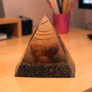 May include: A clear pyramid-shaped orgone energy generator with a copper coil at the top. The pyramid contains dark, organic material and is set on a wooden surface. The pyramid is a decorative item.