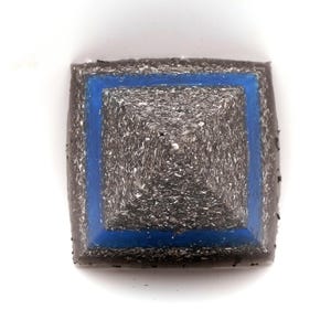 Peut inclure: Objet carr&eacute; en forme de pyramide avec une surface textur&eacute;e gris fonc&eacute;. Un carr&eacute; bleu vif souligne la section centrale de la pyramide. L'objet est pos&eacute; sur un fond blanc, soulignant sa forme g&eacute;om&eacute;trique.
