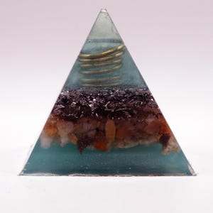May include: A teal-coloured pyramid-shaped resin art piece. It contains layers of dark and light-coloured stones and a coiled gold-coloured wire. The pyramid is set against a white background, showcasing its geometric form.