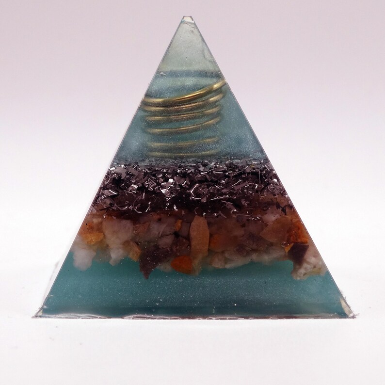 May include: A teal-coloured pyramid-shaped resin art piece. It contains layers of dark and light-coloured stones and a coiled gold-coloured wire. The pyramid is set against a white background, showcasing its geometric form.