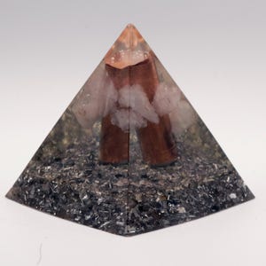 Op de afbeelding: Een piramidevormig decoratief object met een heldere hars buitenkant. Binnenin bevinden zich bruine cilindrische elementen, witte kristallen en een basis van donkere, metaalachtige fragmenten. De top van de piramide is lichtoranje. Ongeveer 8 cm hoog.