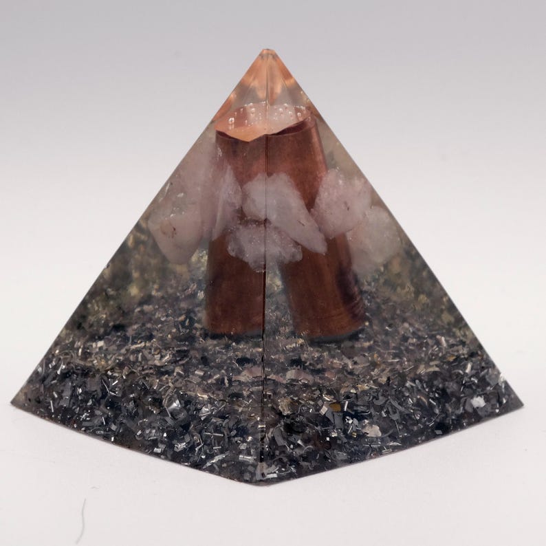Op de afbeelding: Een piramidevormig decoratief object met een heldere hars buitenkant. Binnenin bevinden zich bruine cilindrische elementen, witte kristallen en een basis van donkere, metaalachtige fragmenten. De top van de piramide is lichtoranje. Ongeveer 8 cm hoog.