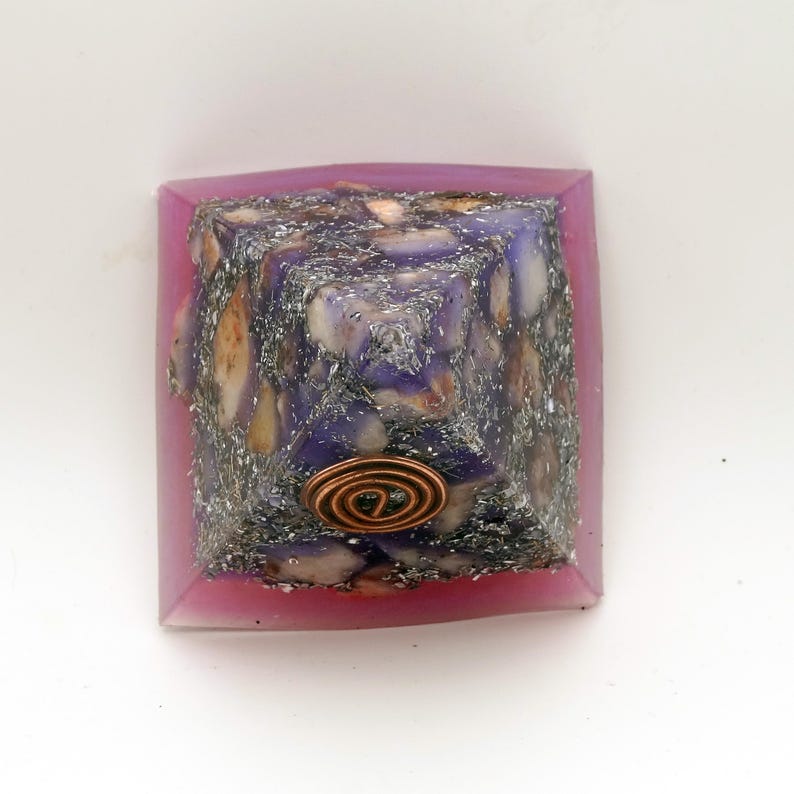 May include: A handcrafted pyramid-shaped object with a copper coil at the base. The pyramid is filled with a mix of purple, white, and tan stones, encased in a pink resin frame. The pyramid is a unique decorative item.