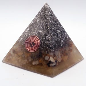 May include: A pyramid-shaped orgone energy generator. The pyramid is made of clear resin with embedded stones and a copper coil. The base is a light brown color, transitioning to a darker gray at the top. The copper coil is in the center.