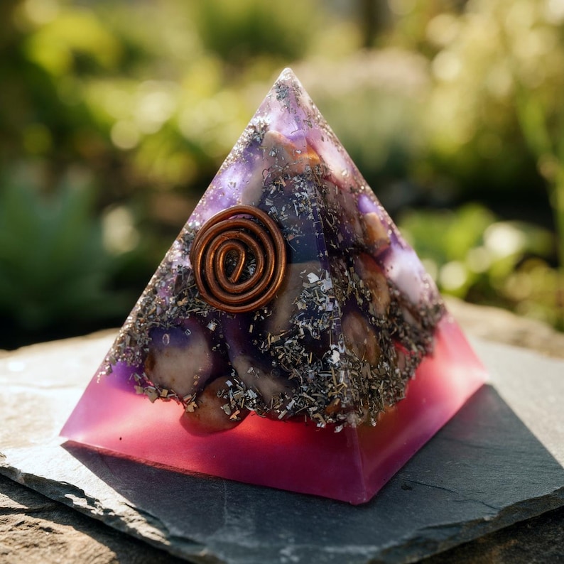 May include: A pyramid-shaped resin sculpture with a copper coil at the top. The pyramid has a pink base that fades into purple, with embedded stones and dried herbs. The pyramid is displayed on a dark gray surface.