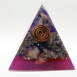 May include: A pyramid-shaped orgone energy generator with a copper coil at the center. The pyramid features layers of purple and magenta resin, with embedded crystals and metallic flakes. The pyramid is a spiritual and decorative item.