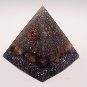 May include: A pyramid-shaped orgone energy generator. The pyramid is made of a clear resin with dark gray and silver inclusions, and small stones. A copper coil is embedded at the top.