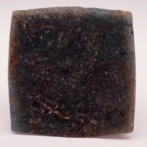 May include: A square, translucent soap bar with a dark, speckled interior. The soap has a blue-green hue around the edges and a dark, almost black center. The texture appears to have embedded particles.