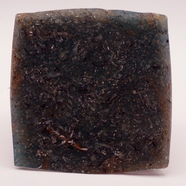May include: A square, translucent soap bar with a dark, speckled interior. The soap has a blue-green hue around the edges and a dark, almost black center. The texture appears to have embedded particles.