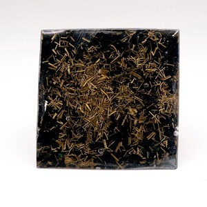 May include: Square, black resin art piece with embedded gold-colored metal shavings. The artwork has a textured appearance with the metallic elements scattered throughout the dark base. The piece is displayed against a white background.