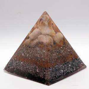 May include: A layered pyramid-shaped object with a clear resin exterior. The pyramid contains various materials, including light-colored stones, dark grey metallic flakes, and a brown layer. The base is a dark, almost black color.