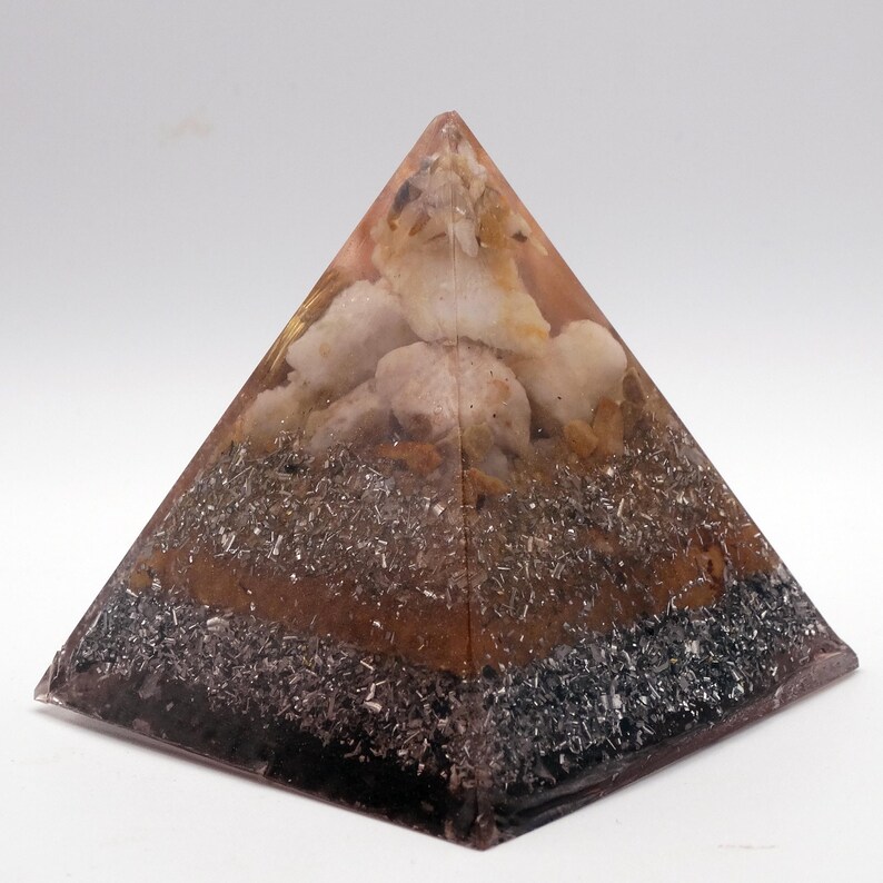 May include: A layered pyramid-shaped object with a clear resin exterior. The pyramid contains various materials, including light-colored stones, dark grey metallic flakes, and a brown layer. The base is a dark, almost black color.