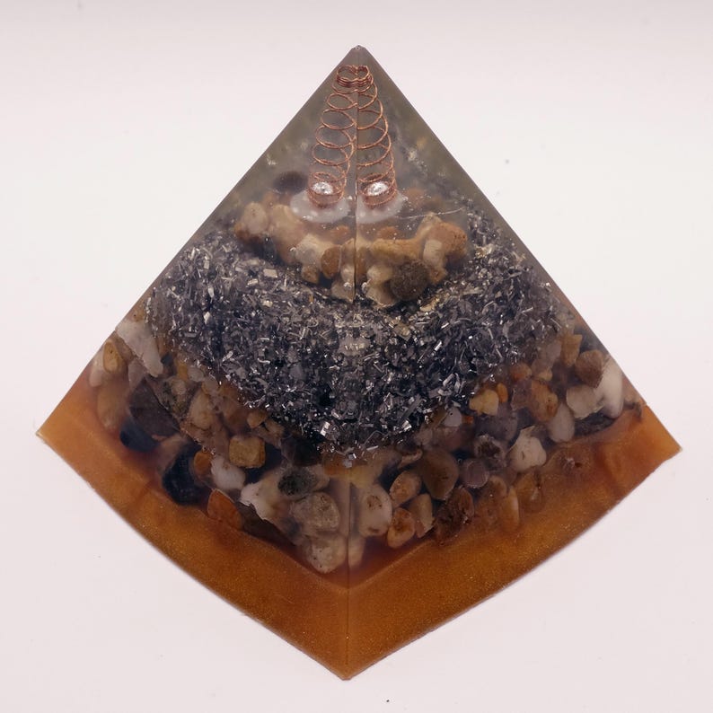 Puede incluir: Un objeto decorativo en forma de pir&aacute;mide con una espiral de cobre en la parte superior. El interior est&aacute; lleno de peque&ntilde;as piedras y escamas met&aacute;licas oscuras, encapsuladas en resina transparente con un borde marr&oacute;n.