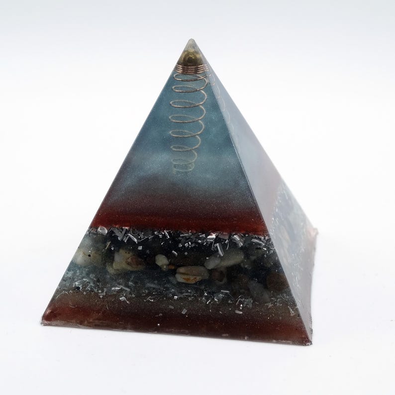 May include: A pyramid-shaped decorative object with a layered design. The top section is a gradient of blue and gray, transitioning to a red band, and a base with embedded stones. A copper coil is at the apex.
