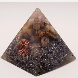 Puede incluir: Una escultura de resina transparente en forma de pir&aacute;mide con piedras incrustadas y una espiral de cobre. La pir&aacute;mide contiene una mezcla de piedras marr&oacute;n claro y blancas, con una base de escamas plateadas. La espiral de cobre est&aacute; centrada.