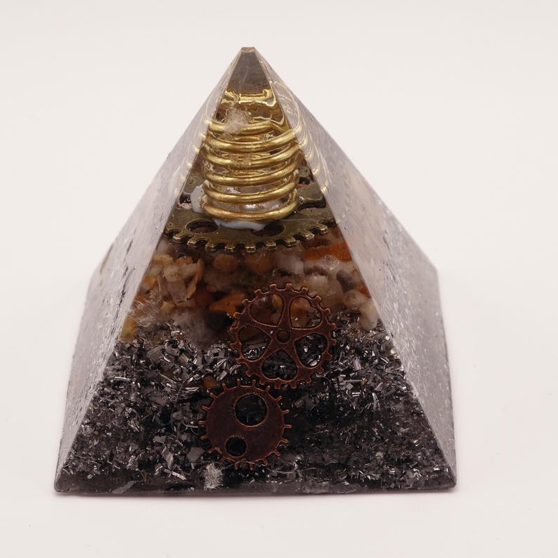 Puede incluir: Un objeto decorativo en forma de pir&aacute;mide con est&eacute;tica steampunk. La pir&aacute;mide est&aacute; hecha de resina transparente, que encierra engranajes, piedras y una bobina dorada. La base es de color gris oscuro con aspecto met&aacute;lico.