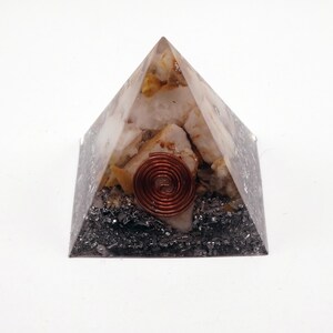May include: A clear pyramid-shaped orgone energy generator. The pyramid contains quartz crystals, a copper coil, and black tourmaline. The copper coil is centered on the pyramid. The base of the pyramid is black.