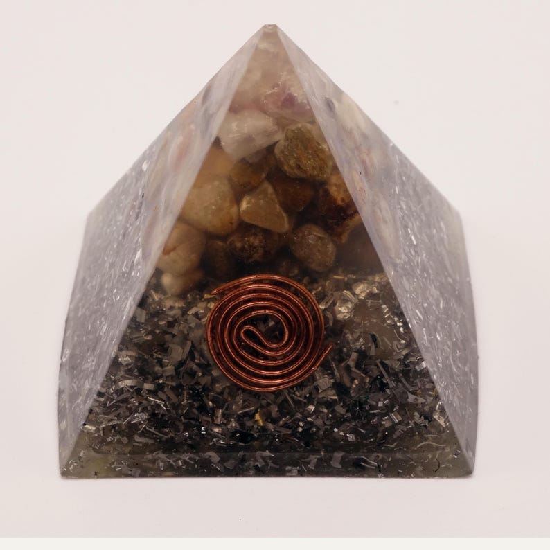 Puede incluir: Un objeto transparente en forma de pir&aacute;mide con una espiral de cobre en la base. La pir&aacute;mide est&aacute; llena de peque&ntilde;as piedras claras y fragmentos cristalinos oscuros. La espiral de cobre es un elemento central.