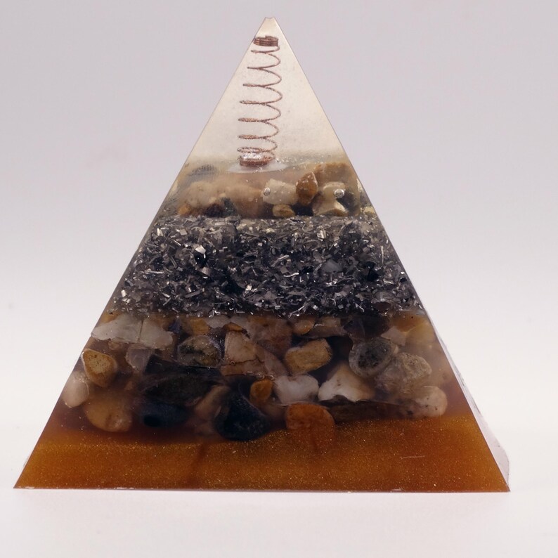 Puede incluir: Una pir&aacute;mide de resina transparente con capas de piedras, minerales y una espiral de cobre en la parte superior. La base es de color marr&oacute;n dorado, y la pir&aacute;mide est&aacute; llena de piedras de varios tonos marrones y grises.
