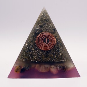 May include: A pyramid-shaped orgone energy generator. The pyramid is clear with a copper coil at the center. The pyramid is filled with dark green and black materials, and a layer of pink resin with small stones at the base.