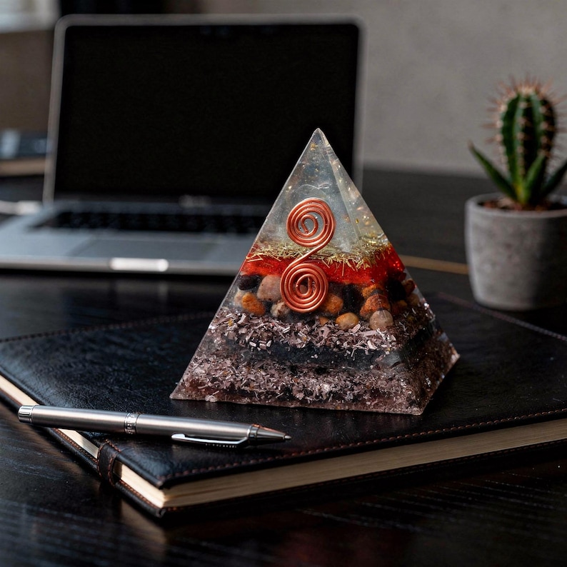 May include: A clear pyramid-shaped resin art piece with copper wire spirals and layers of red, gold, and brown materials. The pyramid is on a black leather-bound notebook with a silver pen. A laptop and a potted cactus are in the background.