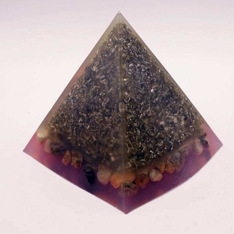 May include: A clear pyramid-shaped resin object with a dark, textured interior. The base is a translucent pink, lined with small, rounded stones. The pyramid's interior appears to contain small, organic materials, creating a unique, decorative piece.