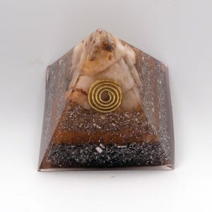 May include: A pyramid-shaped orgone energy generator. The pyramid features a mix of brown, white, and black materials, with a gold-colored spiral at the center. The pyramid is made of resin and is designed to balance energy.