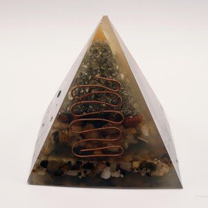 May include: A clear pyramid-shaped resin object with a copper wire coil and embedded stones and minerals. The pyramid has a layered appearance with a mix of colors, including brown, green, and beige. The copper wire is a prominent design element.
