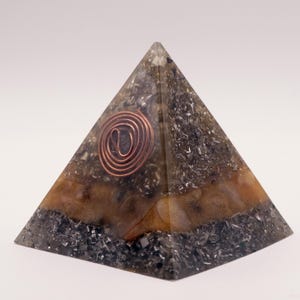 May include: A pyramid-shaped orgone energy generator. The pyramid features a copper coil at the top and is filled with a mix of dark gray and brown materials. The base is a darker shade, with a lighter brown band in the middle.