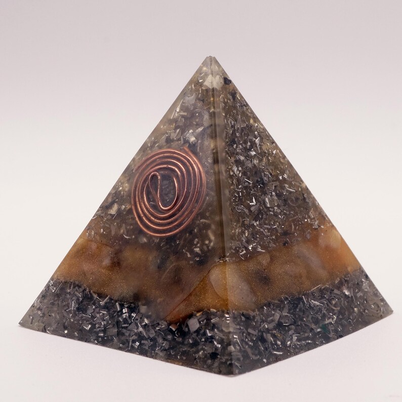 May include: A pyramid-shaped orgone energy generator. The pyramid features a copper coil at the top and is filled with a mix of dark gray and brown materials. The base is a darker shade, with a lighter brown band in the middle.